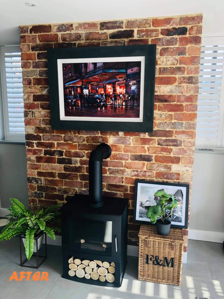 A chimney breast clad in reclaimed brick slips with a cast iron modern log burner in front. There is a picture hanging on the chimney breast.